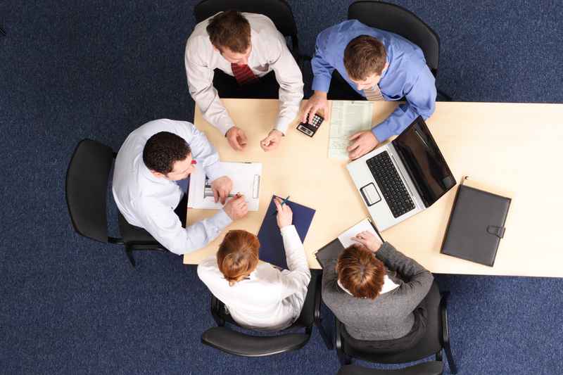 Business professionals gathered around a table reviewing documents and building strategy, working collaboratively in a modern office environment with a positive, focused atmosphere